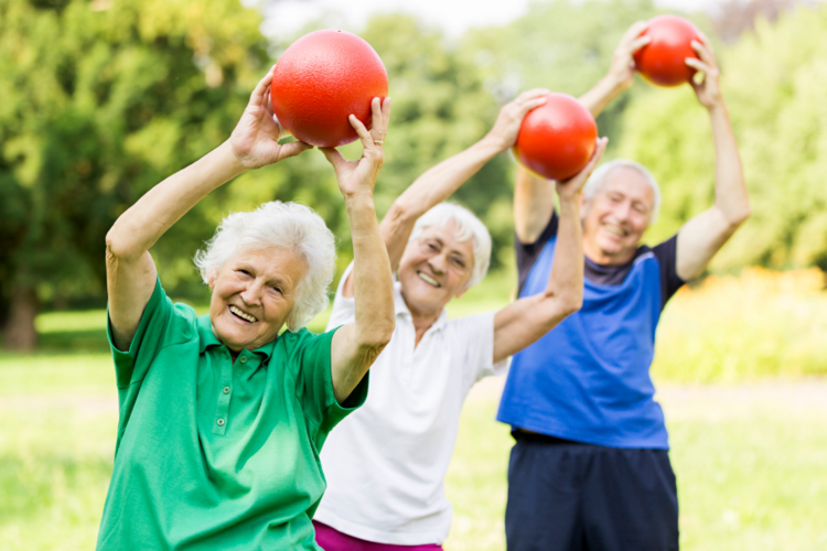 Auf diesem Bild sind drei ältere Personen zu sehen, die jeweils einen roten Ball über den Kopf halten.