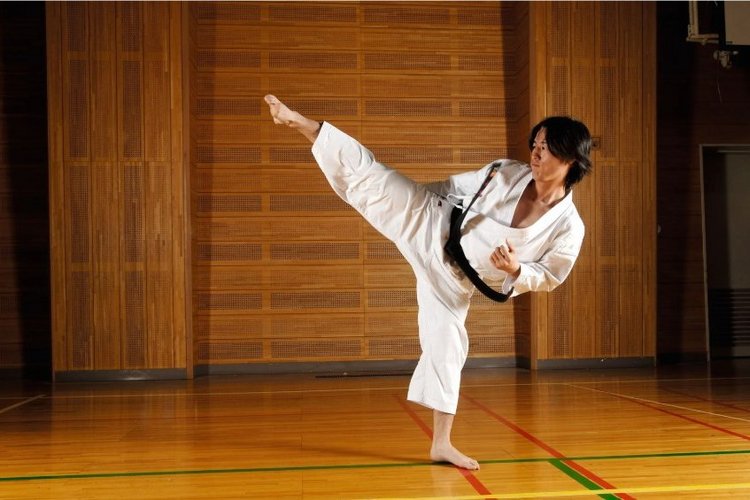 Auf diesem Bild ist eine Person in einem weißen Anzug des Kampfsportes mit schwarzem Gürtel zu sehen. Die Person kickt ein Bein in die Luft. 