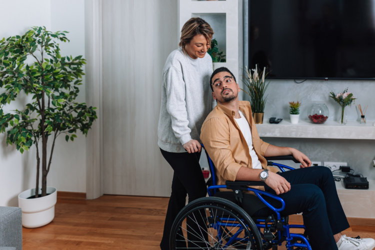 Auf dem Bild ist eine Person im Rollstuhl zu sehen, die von einer anderen geschoben wird. 
