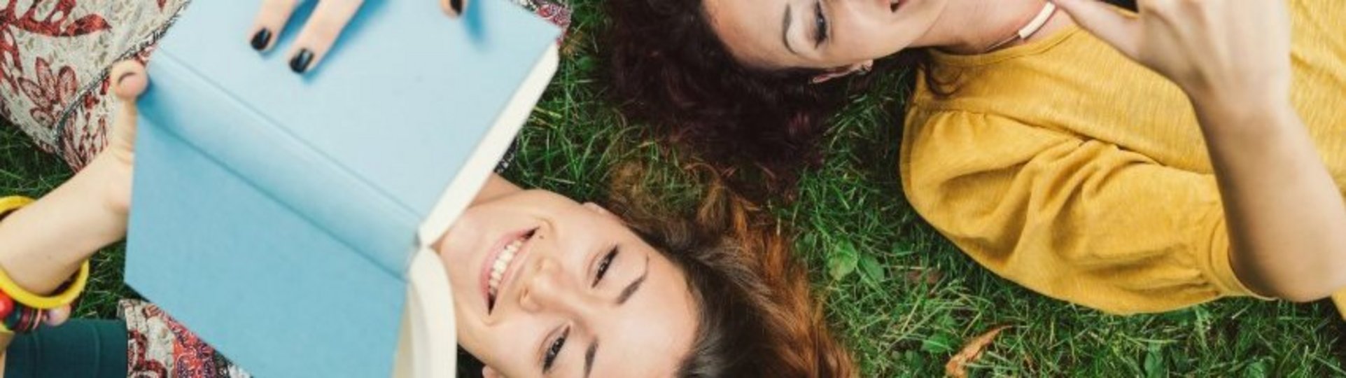 Auf diesem Bild liegen zwei Frauen im Gras. Eine lies ein Buch, die andere guckt in ihr Handy.