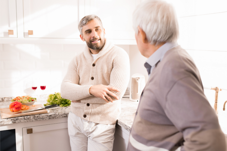 Auf diesem Bild sind ein Mann im mittleren Alter und ein älterer Mann gemeinsam in einer Küche zu sehen. Die beiden Männer unterhalten sich.