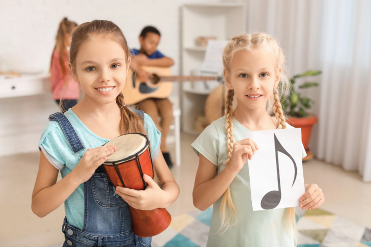 Auf diesem Bild sind zwei Mädchen zu sehen. Das eine Mädchen hält eine Trommel, das andere Mädchen hält einen Zettel mit einer Note.
